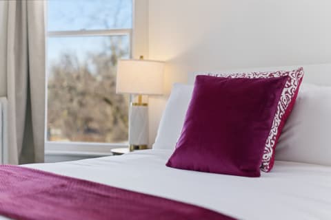 Elegant bedroom with white linens, a purple pillow, and a stylish lamp by the window.