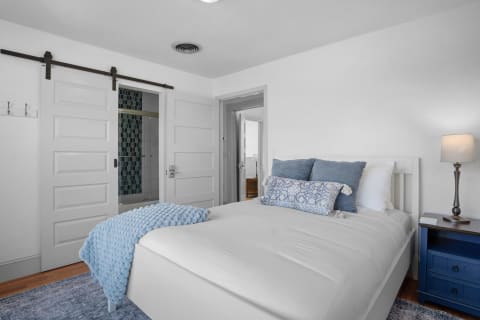 A serene bedroom with white walls and soft blue accents, featuring a neatly made bed, a navy blue nightstand with a lamp, and barn door style sliding doors leading to a bathroom.