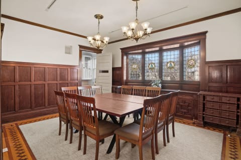A spacious dining room featuring an elegant wooden table surrounded by chairs, with beautiful stained glass windows and warm wood paneling.