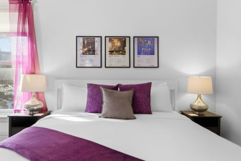 Stylish bedroom with white linens, purple accents, and framed artwork focused on Prince's albums.