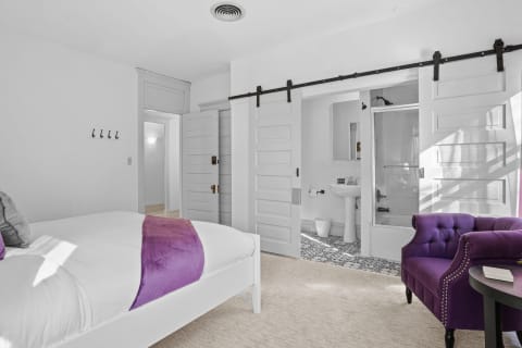 Stylish hotel suite with white bed and purple seating, featuring a barndoor leading to a modern bathroom.