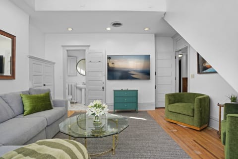 A stylish living room featuring a gray sofa, green armchairs, and a glass coffee table, with a teal cabinet and flat-screen TV.