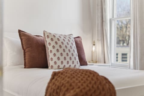 A cozy bedroom featuring soft white linens, decorative pillows, and a warm knitted throw, bathed in natural light.