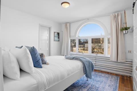 A cozy bedroom featuring white linens, blue accents, and a large arched window with a view outside.