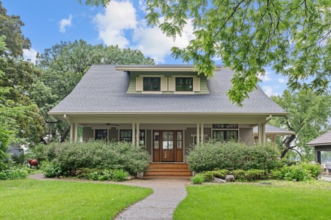 A picturesque two-story house with a gray roof and green shutters, surrounded by greenery.