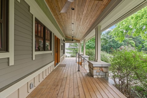 An inviting porch featuring wooden flooring, large windows, and lush greenery.
