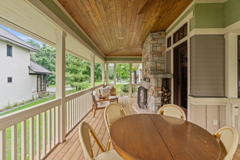 A spacious porch area with a wooden table, chairs, and a stone fireplace, set in a lush green environment.