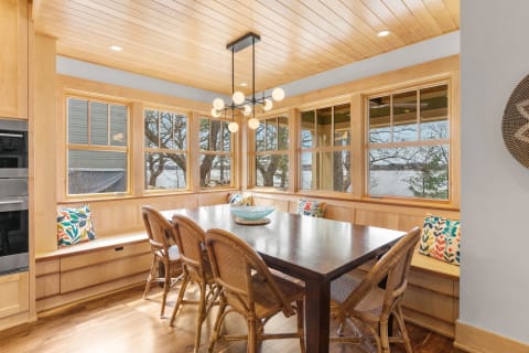 A stylish dining room with wooden elements, a long table, rattan chairs, and large windows displaying trees and water.
