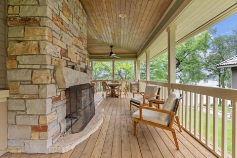 A comfortable porch featuring a stone fireplace and wooden seating, with a view of green trees and water in the background.