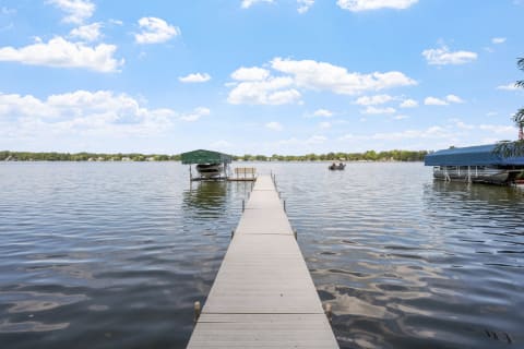 A long dock leads into a calm lake with boats on both sides and a sunny blue sky overhead.