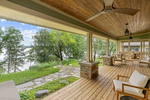 A spacious porch with wooden flooring and ceiling, featuring comfortable chairs and a view of a lake and trees.