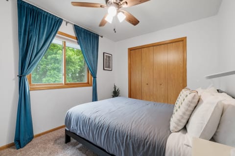 A cozy bedroom featuring a queen bed with a light blue-gray comforter, blue drapes, and a wooden closet.