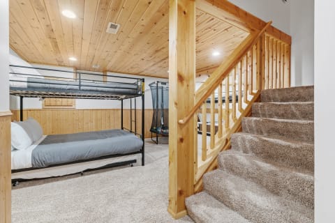 A basement bedroom featuring a black metal bunk bed, colorful toy area, and wooden stairs.