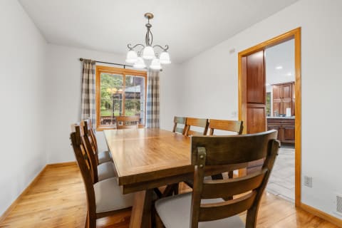 A well-lit dining room with a wooden table, chairs, and a chandelier, showcasing an adjoining kitchen area.