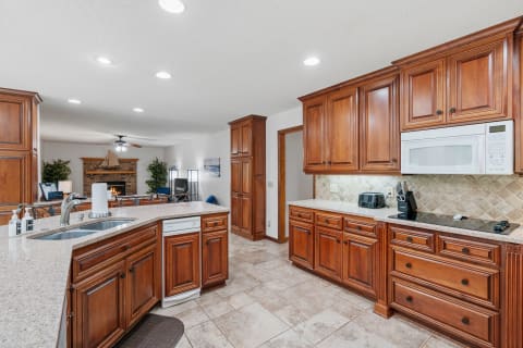 A spacious kitchen featuring wooden cabinetry, granite countertops, and a view of a living room with a fireplace.