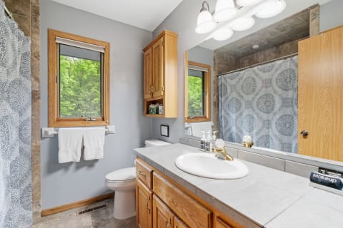 A modern bathroom with a gray color scheme, featuring a sink, wooden cabinets, and a lush green view through the window.
