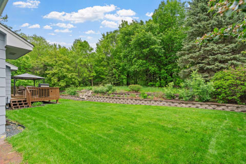 Backyard with a wooden deck, green lawn, and lush trees under a blue sky.