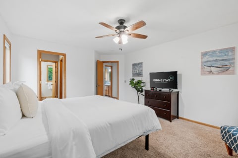 Bright bedroom featuring a neatly made bed, a ceiling fan, a TV, and decorative artwork.