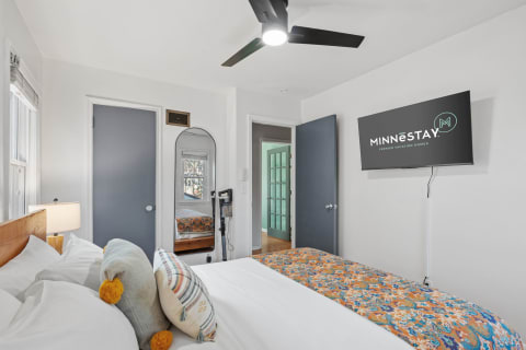 A brightly lit bedroom featuring a comfortable bed with colorful pillows and a mounted TV.