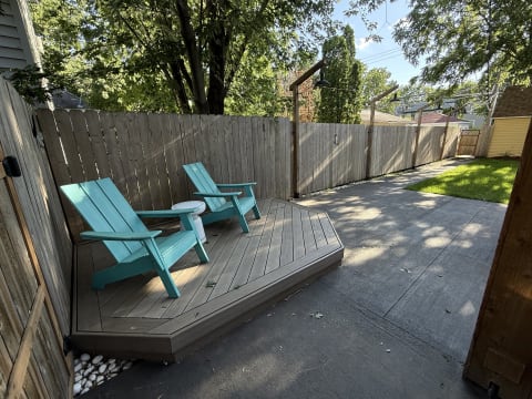 Private backyard deck with turquoise Adirondack chairs and a small white table, surrounded by a wooden fence and greenery.