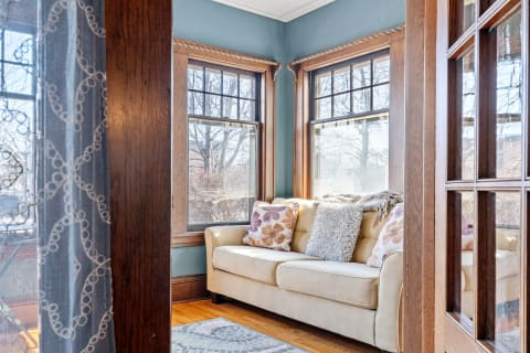 A comfortable reading nook with a beige couch, colorful cushions, and large windows letting in sunlight.