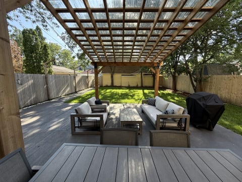 A cozy patio area with comfortable seating beneath a wooden pergola, surrounded by greenery and a barbecue grill.