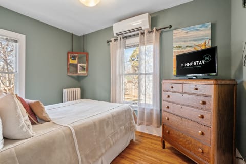 Cozy bedroom with sage green walls, a made bed, wooden dresser, and a view of trees outside the window.