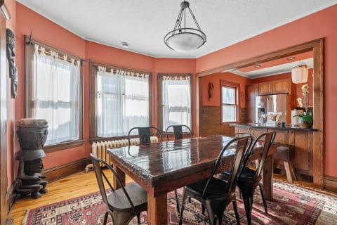 A dining space featuring a large wooden table surrounded by black chairs, with terracotta walls and a view of an adjacent kitchen.