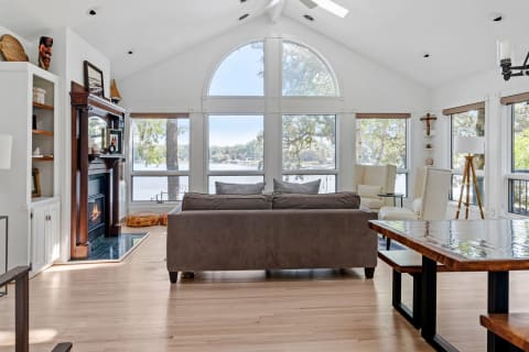 A spacious living room featuring a gray sofa, large windows overlooking a lake, and tasteful wooden decor.