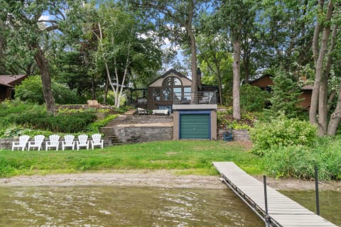 Lakeside view of a modern home with greenery, a dock, and white chairs.