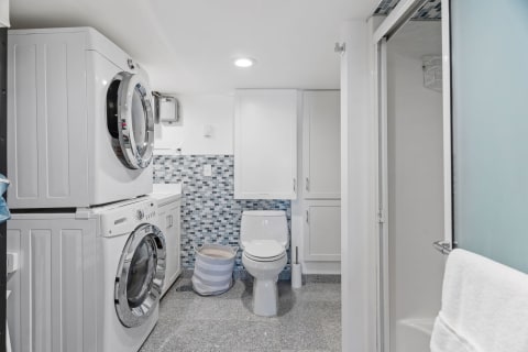 A compact laundry and bathroom area with a stacked washer and dryer, a white toilet, and a mosaic tile backsplash.
