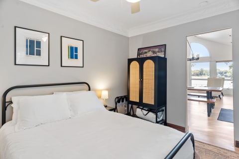 Cozy bedroom with a bed, artwork on the wall, a cabinet, and an open view to another space.