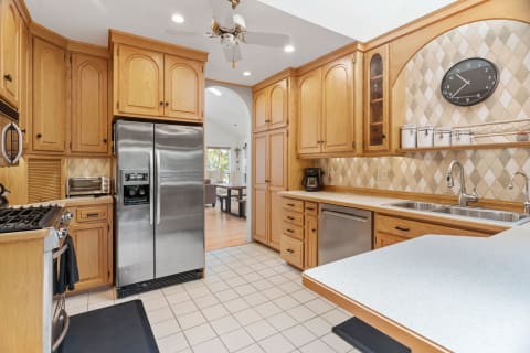 Bright kitchen with wooden cabinets, stainless steel refrigerator, and a dining area visible in the background.