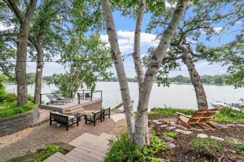 Lakeside patio view with seating and lounge chairs under trees.