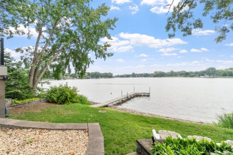 Lakeside view featuring a dock, green grass, and trees under a blue sky with clouds.