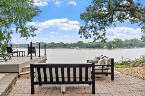 Outdoor seating area overlooking a calm river with trees and blue sky in the background.