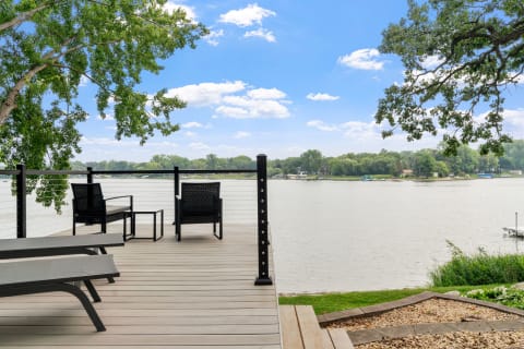 A peaceful waterfront scene featuring two wicker chairs on a deck, looking out over a river with trees and houses in the background.