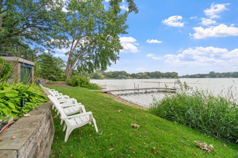 Lakeside scene with white lounge chairs on greenery, a wooden dock, and blue skies.