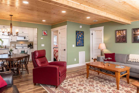 A cozy living room featuring a gray sofa, red recliner, and wooden coffee table on patterned rug, with a warm wood ceiling and a glimpse of the kitchen area.