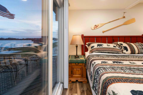 A rustic bedroom with red bed frame, patterned bedding, a lamp, and a view of the lake through the window.