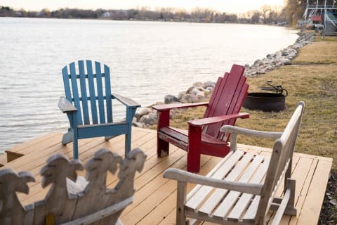 Three wooden chairs, one blue, one red, and one white, arranged by a serene lake on a wooden deck.