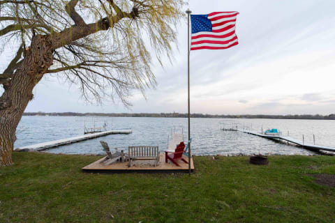 Wooden deck with Adirondack chairs by a lake under a cloudy sky.