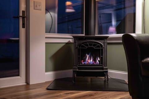 A gas stove with flickering flames in a corner of a cozy living room, surrounded by walls painted in a soothing green tone and illuminated by indoor lights.