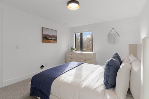 A cozy modern bedroom featuring a bed with blue pillows, a dresser, and a window with outdoor views.