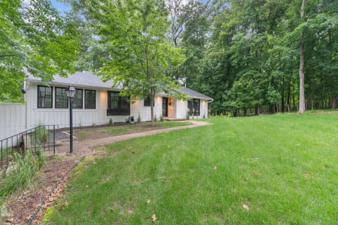 A single-story house with white siding and a brick pathway leading to the entrance, set in a lush green yard.
