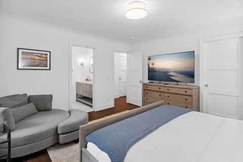 A modern bedroom with white walls, a gray sofa, and a flat-screen TV showing a beach scene.