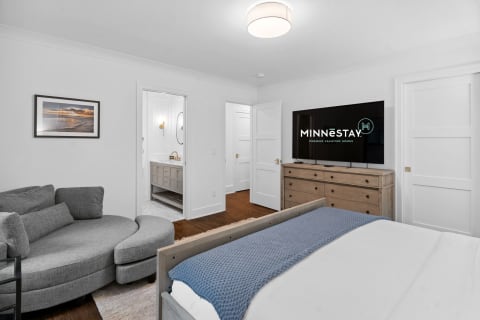 A stylish bedroom with a gray sectional sofa, a wooden dresser with a TV, and a modern bathroom doorway.