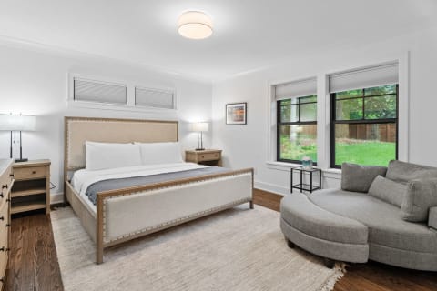 Modern bedroom with a beige bed, gray sectional, and large windows overlooking a green backyard.