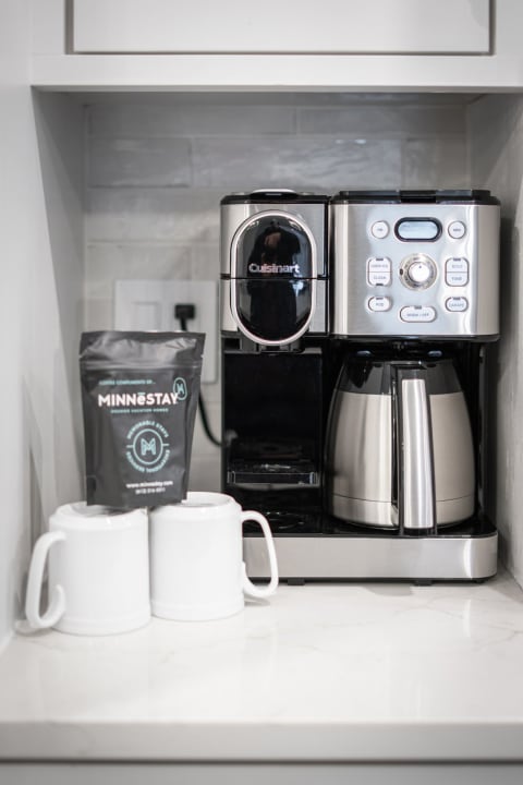 A Cuisinart coffee maker next to two white mugs and a black coffee package on a kitchen countertop.