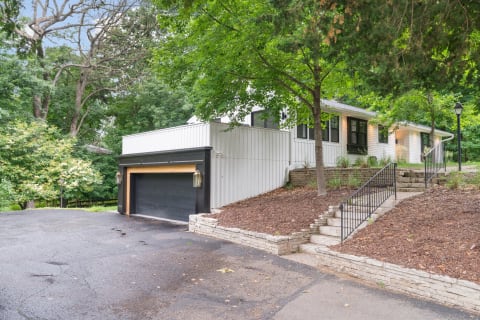 A white house with large windows, a black garage, and scenic landscaping surrounded by trees.